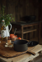 Load image into Gallery viewer, Settle | Netherton Foundry - mid-view of a black spun iron stock pot set on an antique wooden chopping board, next to its lid, chopped onions with their skins, vine tomatoes, a bottle of olive oil, fresh herbs in an old enamel jug,  in the heritage kitchen of one of Settle's luxury retreats.