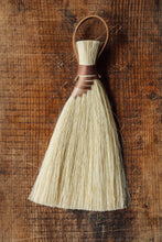 Load image into Gallery viewer, SETTLE | Wild Crafted Home Handmade Hand Brush - a triangular shaped handmade brush with pale gold fibres and a stem made of wound pale and golden fibres with a looped leather band, placed on antique timbers at Settle.