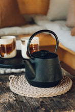Load image into Gallery viewer, Shop at Settle The Chemists Daughter - a circle-woven trivet, handmade from 100% recycled pale cotton, in action with a black iron teapot placed on it, next to two handmade ceramic mugs. Tea time!