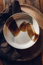 Load image into Gallery viewer, Settle | By Noo Ceramics Brushstroke Open Bowl - close overview of a pale handmade ceramic bowl with a generous base and gently curved sides, hand decorated with a thick brush stroke of chocolate brown across the bowl's interior. Placed on a broad wooden plate next to a dark brown linen napkin, all sitting on an antique wooden stool on antique timbers at Settle.