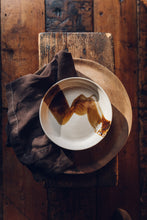 Load image into Gallery viewer, Settle | By Noo Ceramics Brushstroke Open Bowl - overview of a pale handmade ceramic bowl with a generous base and gently curved sides, hand decorated with a thick brush stroke of chocolate brown across the bowl's interior. Placed on a broad wooden plate next to a dark brown linen napkin, all sitting on an antique wooden stool on antique timbers at Settle.
