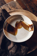Load image into Gallery viewer, Settle | By Noo Ceramics Brushstroke Open Bowl - close view of a pale handmade ceramic bowl with a generous base and gently curved sides, hand decorated with a thick brush stroke of chocolate brown across the bowl's interior. Placed on a broad wooden plate next to a dark brown linen napkin, all sitting on an antique wooden stool on antique timbers at Settle.