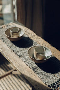 Shop with SETTLE | Cabin Woven - two handmade ceramic bowls are set on a handwoven table runner made from handspun wool, with tasselled ends and subtle brown on charcoal stripe, on an antique timber trestle at Settle. 