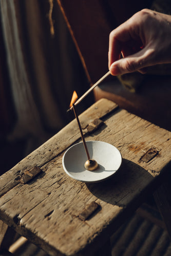 Shop with SETTLE | Essence+Alchemy Autumna Incense Sticks - a hand is in frame lighting an incense stick with a lighted match; the incense stick is held in a small golden dome holder within a pale stoneware bowl, all placed on an antique wooden side table in one of Settle's luxury retreats.