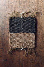 Load image into Gallery viewer, Shop at Settle | Cabin Woven ~ colour photograph of a handwoven rectangular placemat in charcoal grey and fawn brown fibres, with fronded ends, set on an antique wooden table.  