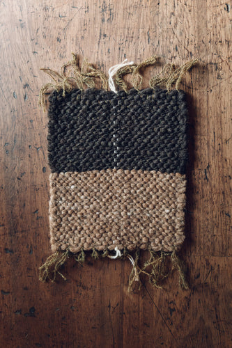 Shop at Settle | Cabin Woven ~ colour photograph of a handwoven rectangular placemat in charcoal grey and fawn brown fibres, with fronded ends, set on an antique wooden table.  