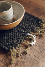 Load image into Gallery viewer, Shop with Settle | Cabin Woven ~ Close-up detail of fronded edge of handwoven placemat in charcoal grey. A hand thrown ceramic piece sits on set on the placement, which is set on an antique wooden table. 