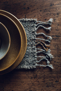 Shop with SETTLE | Cabin Woven - a place setting of handmade ceramics is set on a handwoven table runner made from handspun wool, with tasselled ends and subtle brown on charcoal stripe, on an antique timber table at Settle. 