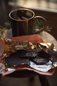 Bar of Pump Street Chocolate's Gingerbread Chocolate snapped into several pieces, lying in its beautiful wrapping with a mug of hot drink and a sprig of pine on an antique timber stool at Settle Norfolk UK | Copyright Settle Norfolk, UK