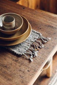 Shop with SETTLE | Cabin Woven - a place setting of handmade ceramics is set on a handwoven table runner made from handspun wool, with tasselled ends and subtle brown on charcoal stripe, on an antique timber table at Settle. 