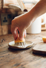 Load image into Gallery viewer, SETTLE | Every Story Ceramics Citrus Juicer - a hand holds half a lemon on a ceramic citrus juicer handmade in natural speckled clay, set on an antique timber kitchen surface with kitchen utensils in the background at Settle.