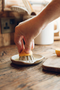 SETTLE | Every Story Ceramics Citrus Juicer - a hand holds half a lemon on a ceramic citrus juicer handmade in natural speckled clay, set on an antique timber kitchen surface with kitchen utensils in the background at Settle.