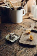 Load image into Gallery viewer, SETTLE | Every Story Ceramics Citrus Juicer - a ceramic citrus juicer handmade in natural speckled clay holds freshly squeezed lemon juice, next to a wooden chopping board with two halves of a lemon and a knife, set on an antique timber kitchen surface with kitchen utensils in the background at Settle.