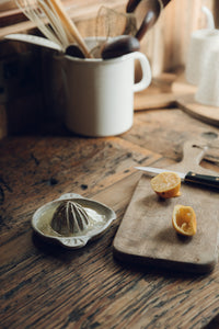 SETTLE | Every Story Ceramics Citrus Juicer - a ceramic citrus juicer handmade in natural speckled clay holds freshly squeezed lemon juice, next to a wooden chopping board with two halves of a lemon and a knife, set on an antique timber kitchen surface with kitchen utensils in the background at Settle.