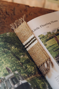 Shop with Settle | Cabin Woven - handwoven from handspun wool bookmark with tasselled ends and central stripe detail; fawn brown with white and charcoal stripe. Shown lying on the open pages of a lifestyle book.