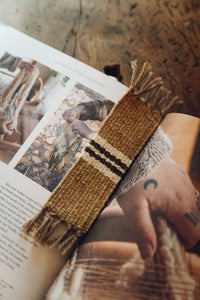 Shop with Settle | Cabin Woven - handwoven bookmark, from handspun wool, with tasselled ends and central stripe detail; fawn brown with white and charcoal stripe. Shown lying on the open pages of a lifestyle book.