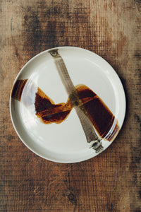 Settle | By Noo Ceramics Brushstroke Dinner Plate - a handmade pale ceramic dinner plate hand decorated with two brushstrokes in dark brown paint, set on antique timbers in one of Settle's luxury retreats.  