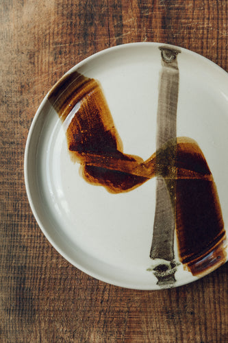 Settle | By Noo Ceramics Brushstroke Dinner Plate - closer view of a handmade pale ceramic dinner plate hand decorated with two brushstrokes in dark brown paint, set on antique timbers in one of Settle's luxury retreats.