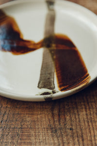 Settle | By Noo Ceramics Brushstroke Dinner Plate - closeup on a handmade pale ceramic dinner plate hand decorated with two brushstrokes in dark brown paint, set on antique timbers in one of Settle's luxury retreats.