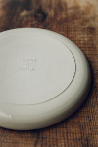 Settle | By Noo Ceramics Brushstroke Dinner Plate - base of a handmade pale ceramic dinner plate, marked with the words 'by.noo. Settle' and Settle's leaf motif, set on antique timbers in one of Settle's luxury retreats.