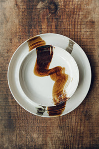 Settle | By Noo Ceramics Brushstroke Dinner Plate - two pieces of handmade pale ceramics from the same collection - a dinner plate beneath an open bowl - hand decorated with two brushstrokes in dark brown paint, set on antique timbers in one of Settle's luxury retreats.