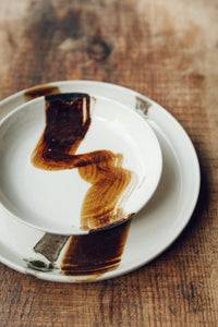 Settle | By Noo Ceramics Brushstroke Open Bowl - close view of two pale handmade ceramics, a side plate and a dinner plate, each hand decorated with a thick brush stroke of chocolate brown across the interior of each piece, set on antique timbers at Settle.  