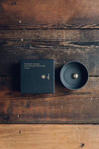 Settle | Ume ~ overview of an elegant black stoneware bowl containing a small gold-coloured incense dome, next to a square black box with Ume's motif and name in gold, all set on antique timbers in one of Settle's luxury retreats. 