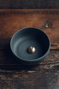 Settle | Ume ~ closer diagonal view of an elegant black stoneware bowl containing a small gold-coloured incense dome, next to a square black box with Ume's motif and name in gold, all set on antique timbers in one of Settle's luxury retreats.