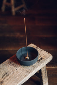 Settle | Ume ~ an elegant black stoneware bowl containing a small gold-coloured incense dome which holds a lighted incense stick, sitting on an antique trestle in one of Settle's luxury retreats.