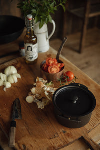 Settle | Netherton Foundry - mid-view of a black spun iron stock pot and its lid set on an antique wooden chopping board alongside chopped onions, a kitchen knife, vine tomatoes, a bottle of olive oil, fresh herbs in an old enamel jug,  in the heritage kitchen of one of Settle's luxury retreats.