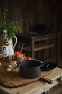 Settle | Netherton Foundry - mid-view of a black spun iron stock pot set on an antique wooden chopping board, next to its lid, chopped onions with their skins, vine tomatoes, a bottle of olive oil, fresh herbs in an old enamel jug,  in the heritage kitchen of one of Settle's luxury retreats.