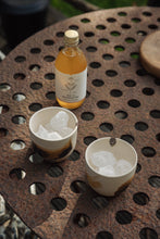 Load image into Gallery viewer, Settle | By Noo Ceramic Brushstroke Collection - two small pale ceramic tumblers, decorated with brush strokes of chocolate brown, are filled with ice and set next to a bottle of botanical drink on a rustic metal surface outdoors on a sunny day at Settle.