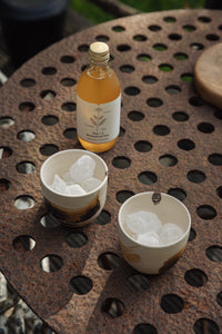 Settle | By Noo Ceramic Brushstroke Collection - two small pale ceramic tumblers, decorated with brush strokes of chocolate brown, are filled with ice and set next to a bottle of botanical drink on a rustic metal surface outdoors on a sunny day at Settle.