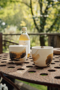 Settle | By Noo Ceramic Brushstroke Collection - two small pale ceramic tumblers, decorated with brush strokes of chocolate brown, are filled with ice and set next to a bottle of botanical drink on a rustic metal surface outdoors on a sunny day at Settle.
