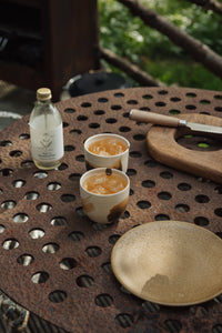 Settle | By Noo Ceramic Brushstroke Collection - two small pale ceramic tumblers, decorated with brush strokes of chocolate brown, are filled with ice and set next to a bottle of botanical drink and other tableware on a rustic metal surface outdoors on a sunny day at Settle.