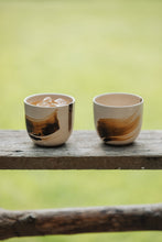 Load image into Gallery viewer, Settle | By Noo Ceramic Brushstroke Collection - two small pale ceramic tumblers, decorated with brush strokes of chocolate brown, are set on a wooden surface outdoors in a sunny green space at Settle. One tumbler contains an iced drink.