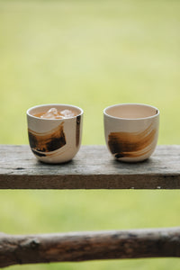 Settle | By Noo Ceramic Brushstroke Collection - two small pale ceramic tumblers, decorated with brush strokes of chocolate brown, are set on a wooden surface outdoors in a sunny green space at Settle. One tumbler contains an iced drink.