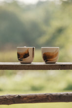 Load image into Gallery viewer, Settle | By Noo Ceramic Brushstroke Collection - two small pale ceramic tumblers, decorated with brush strokes of chocolate brown, are set on a wooden surface outdoors in a sunny green space at Settle.