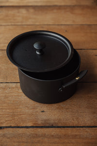 Settle | Netherton Foundry - closer view of a black spun iron stock pot and its lid set on antique wooden timbers.