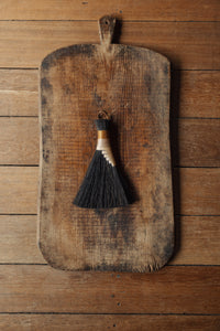 SETTLE | Wild Crafted Home Handmade Hand Brush - a triangular shaped handmade brush with dark brown fibres and a stem made of wound pale and golden fibres with a looped leather band, placed on a large wooden chopping board on a contemporary planked surface at Settle.