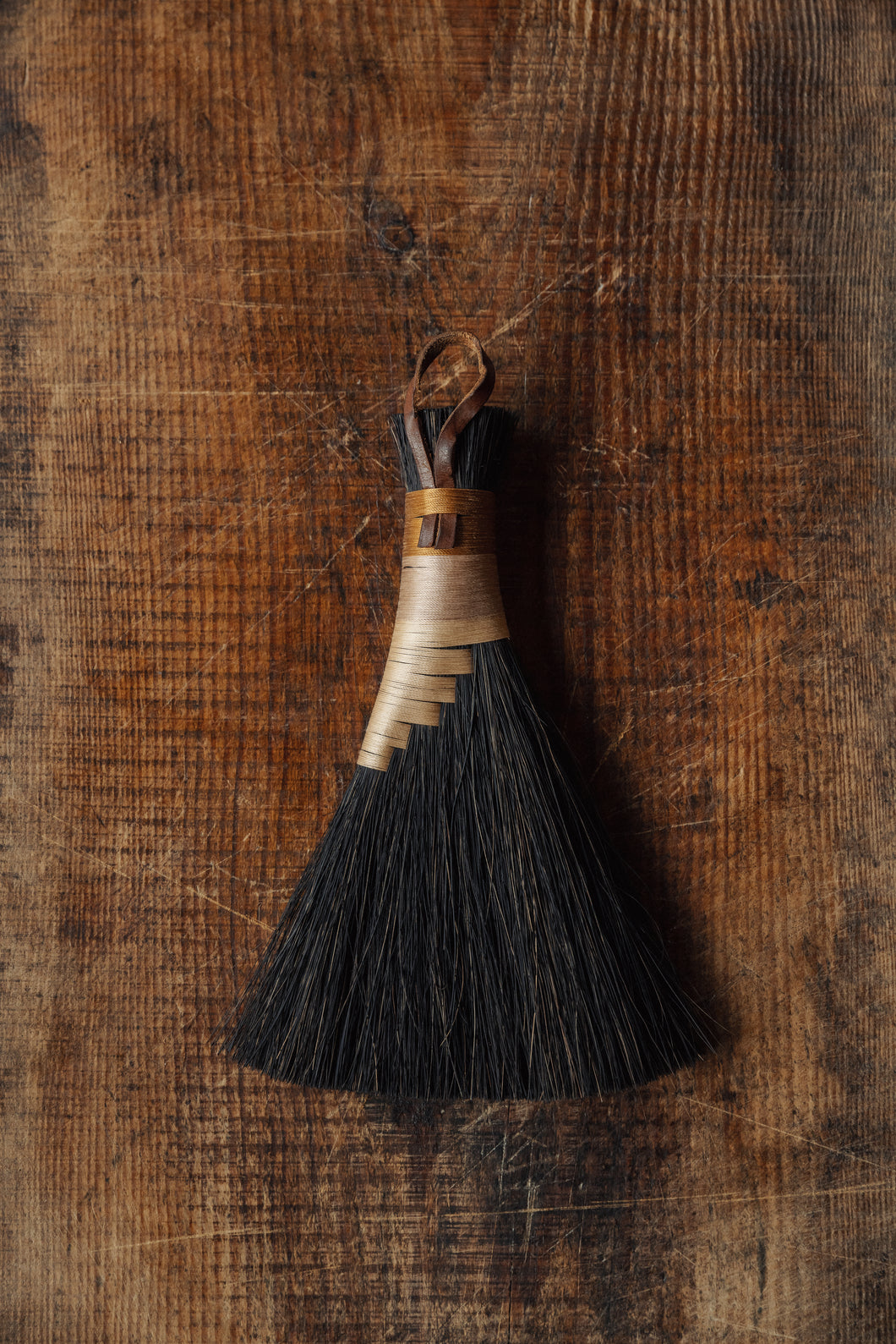 SETTLE | Wild Crafted Home Handmade Hand Brush - a triangular shaped handmade brush with dark brown fibres and a stem made of wound pale and golden fibres with a looped leather band, placed on antique timbers at Settle.