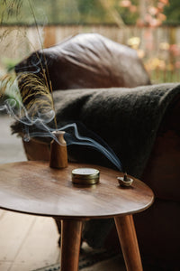 Shop with Settle | Essence + Alchemy Hiems Incense Cones - a lighted incense cone in a gleaming incense dome on a pale ceramic dish set on a contemporary wooden side table at Settle.