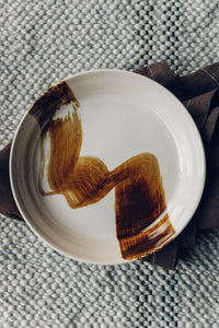 Settle | By Noo Ceramics Brushstroke Open Bowl - close view of a pale handmade ceramic bowl with a generous base and gently curved sides, hand decorated with a thick brush stroke of chocolate brown across the bowl's interior. Set on a dark brown linen napkin, upon a pale green woven textile.  