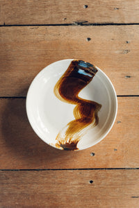 Settle | By Noo Ceramics Brushstroke Open Bowl - over view of a pale handmade ceramic bowl with a generous base and gently curved sides, hand decorated with a thick brush stroke of chocolate brown across the bowl's interior, set on mid-brown aged timbers.  
