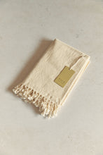 Load image into Gallery viewer, SETTLE | Pico Goods Hand Towel - handwoven organic cotton hand towel in a natural off-white with tasselled edging and a Pico Goods label against a soft peach marble background at Settle.