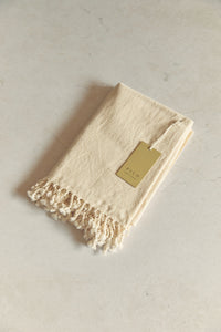 SETTLE | Pico Goods Hand Towel - handwoven organic cotton hand towel in a natural off-white with tasselled edging and a Pico Goods label against a soft peach marble background at Settle.