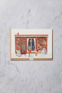 Shop at Settle | Harriet Watson - charming hand illustration , in colour, of one of Settle's repurposed railway carriage stays looking festive with a wreath on the door, lighted candles. In front on its terrace two white people are toasting marshmallows over a lighted fire pit, and next to the a table is set with a candle, drinks, and a blanket slung over the back of a chair. The card is set against a pale grey marble background.