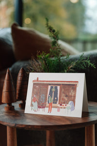 Shop at Settle | Harriet Watson - charming hand illustration , in colour, of one of Settle's repurposed railway carriage stays looking festive with a wreath on the door, lighted candles. In front on its terrace two white people are toasting marshmallows over a lighted fire pit, and next to the a table is set with a candle, drinks, and a blanket slung over the back of a chair. In the interior of Settle's luxurious Cabin.    