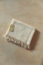 Load image into Gallery viewer, SETTLE | Pico Goods Bath Towel - handwoven organic cotton bath towel in a natural off-white with tasselled edging and a Pico Goods label against a soft peach marble background at Settle.