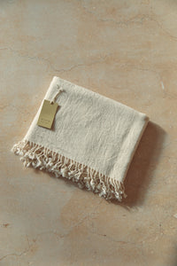 SETTLE | Pico Goods Bath Towel - handwoven organic cotton bath towel in a natural off-white with tasselled edging and a Pico Goods label against a soft peach marble background at Settle.